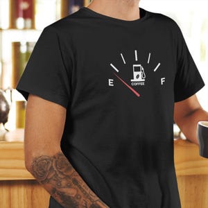 Könnte beinhalten: Schwarzes T-Shirt mit einer weißen Grafik einer Tankanzeige mit einer Kaffeebohne in der Tankpumpe. Die Anzeige zeigt "E" für leer und "F" für voll an. Der Zeiger zeigt auf "E". Der Text "COFFEE" befindet sich unter der Tankpumpe.