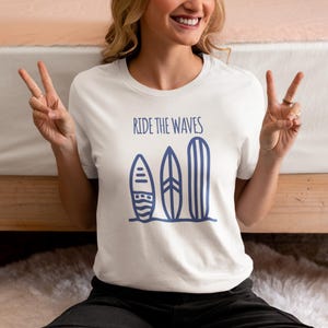Puede incluir: Camiseta blanca con la frase "Ride the Waves" sobre tres tablas de surf azules. Las tablas de surf son de diferentes formas y tamaños. La camiseta es de un material suave.