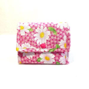 May include: A small fabric wallet featuring a pink background with a repeating pattern of white daisies with yellow centers and green leaves. The wallet has a rectangular flap secured by a single bright pink circular snap button closure.