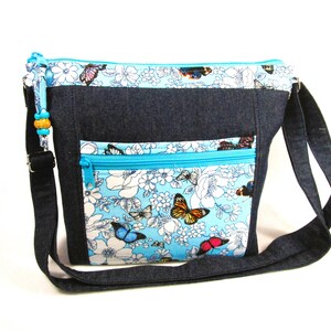 May include: A denim crossbody bag with a light blue floral and butterfly print on the front pocket and top panel. The bag has a turquoise zipper and a black adjustable strap. The bag has a beaded charm on the zipper.