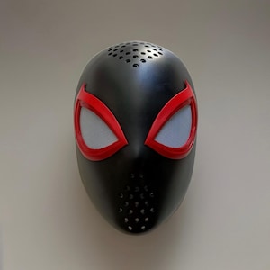 May include: A black mask with red eye frames and white mesh eye coverings. The mask has small ventilation holes at the top and bottom. This mask is inspired by a superhero.