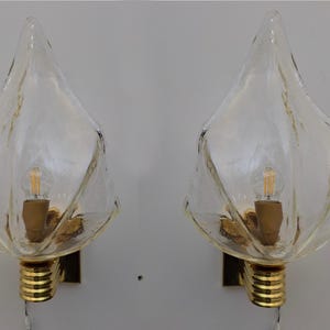 May include: Two clear glass wall sconces with a textured, petal-like design. Each sconce features a gold-colored base and a visible lightbulb. The sconces are mounted on a gold-colored square base, creating a vintage aesthetic.