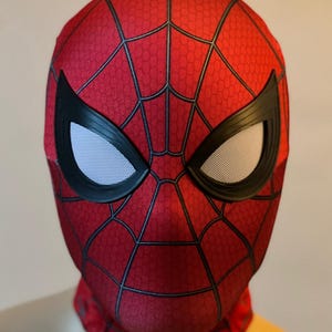 May include: A red and black Spider-Man mask with a web pattern. The mask features black eye frames with white mesh eye coverings. The mask is made of a textured fabric and covers the entire head and neck.