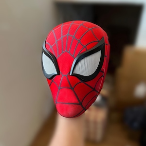 Pode incluir: Uma máscara vermelha do Homem-Aranha com teias pretas e contornos dos olhos. As aberturas dos olhos são brancas com uma textura de malha. A máscara é de cor vermelha vibrante, e as linhas pretas criam um padrão de teia na superfície.