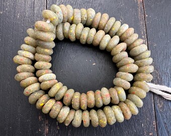 May include: Strand of olive-green, oval-shaped beads with speckled red and yellow accents. The beads are strung on a natural-colored cord, creating a necklace or bracelet component. The beads are displayed on a dark wooden surface.