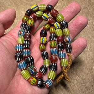 Puede incluir: Un collar de cuentas coloridas en forma de barril, en tonos amarillo, verde, rojo, azul y negro. Las cuentas están ensartadas en un cordón marrón, ideal como collar o elemento decorativo.