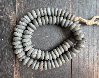 May include: A strand of gray, oval-shaped beads with a rough texture. The beads are strung together with a natural-colored twine. The beads have small yellow and brown accents. The beads are displayed on a dark wooden surface.