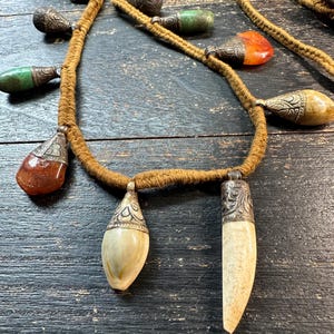 Può includere: Una lunga collana con un cordino spesso avvolto in fibra marrone, decorata con molteplici pendenti. I pendenti includono pietre lisce e levigate nei toni dell'arancione, del verde e della crema, insieme a tappi in metallo scuro inciso e forme intagliate.