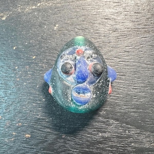 May include: A colorful glass sculpture of a face with black eyes, a blue nose and mouth, and red accents. The sculpture has a green and blue body and is set against a dark wooden surface.