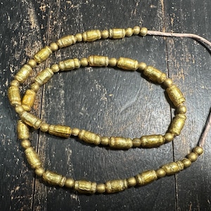 Puede incluir: Un collar de cuentas doradas con una mezcla de formas cilíndricas y esféricas ensartadas en un cordón fino de color claro. Las cuentas cilíndricas tienen un acabado texturizado y desgastado, mientras que las cuentas esféricas pequeñas son lisas.