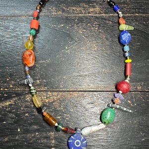 May include: A multi-coloured beaded necklace featuring an assortment of glass, stone, and ceramic beads in various shapes and sizes. The necklace includes a prominent blue circular pendant with white radial lines and a central white circle.