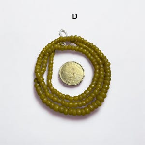Pu&ograve; includere: Collana di perline verde oliva con una moneta. La collana &egrave; composta da piccole perline rotonde. La moneta &egrave; color oro e ha del testo sopra. La collana &egrave; avvolta a forma circolare.