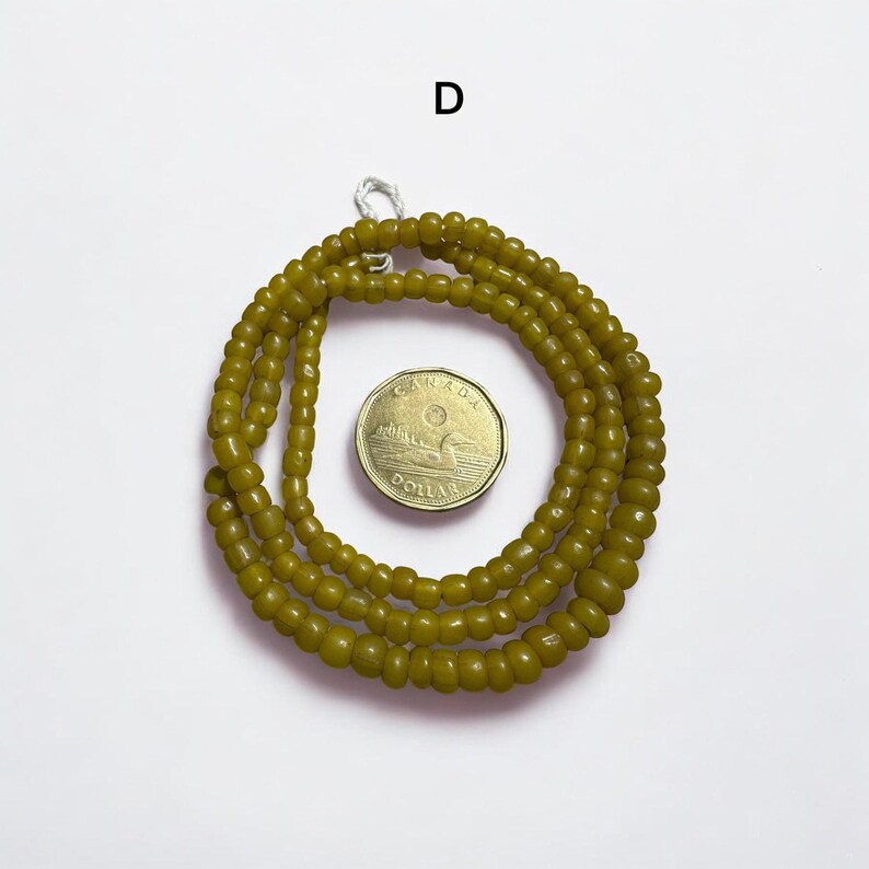 Pu&ograve; includere: Collana di perline verde oliva con una moneta. La collana &egrave; composta da piccole perline rotonde. La moneta &egrave; color oro e ha del testo sopra. La collana &egrave; avvolta a forma circolare.