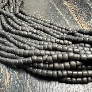 Puede incluir: Una colección de múltiples hilos de pequeñas cuentas de color negro mate dispuestas en una formación curva. Cada cuenta individual presenta una forma redondeada ligeramente irregular con un color gris carbón consistente y una textura suave.