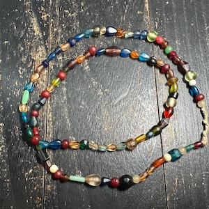 May include: A long strand of multi-coloured glass beads featuring a variety of shapes, sizes, and translucent tones including red, blue, green, yellow, and brown. The beads are strung on a thin cord with a knotted end, creating a continuous loop.