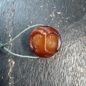May include: A small, round, reddish-brown carved stone bead with an owl design. The owl has large eyes and a detailed feather pattern. A green string is attached to the bead, suggesting it could be used as a pendant or charm.