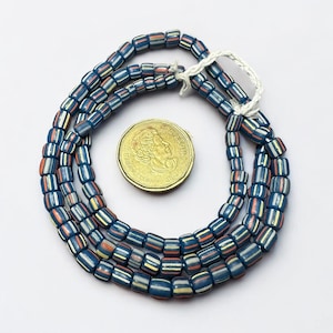 Puede incluir: Hebras de cuentas a rayas azules, rojas, amarillas y blancas. Las cuentas son rectangulares y están ensartadas. Una moneda de oro está al lado de las cuentas.
