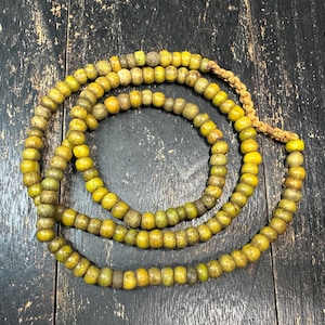 May include: A long strand of round, weathered yellow beads with a matte, textured finish. The beads are strung together on a light brown braided cord, forming a circular loop with a visible knotted section at the top of the strand.