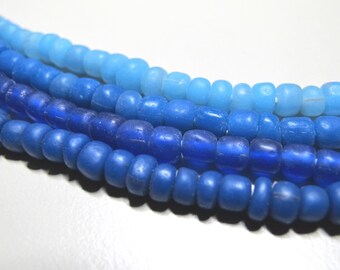 May include: Close-up of four strands of blue glass beads. The beads vary in shade from light to dark blue. The beads are round and have a matte finish. The strands are arranged in parallel lines.