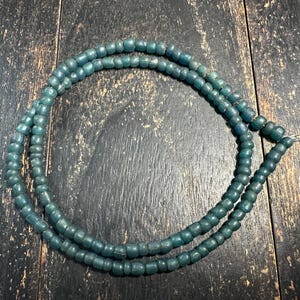 May include: A strand of small, round, teal-colored beads arranged in a circular shape. The beads have a matte, slightly irregular texture and are strung together on a thin, metallic wire that is visible at the end of the bead strand.