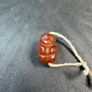 May include: A small, carved, reddish-brown figurine of a seated figure with a string attached. The figurine has a rounded head, a defined face, and a segmented body. The string is a light beige color.