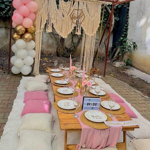 May include: A low wooden picnic table set for a party with white plates, woven placemats, pink table runners, and cushions. A light box displays the word BRIDE, and a macrame backdrop hangs behind the table with a balloon arch in pink, gold, and white.