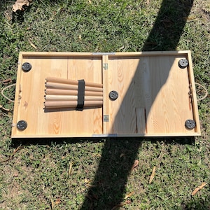 May include: A rectangular light-coloured wooden folding table with a natural grain finish. The underside features four black circular feet at the corners, a central black circular locking mechanism, and five detachable wooden legs secured by a black strap.