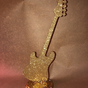 以下が含まれることがあります： 丸い台座が付いた金色のキラキラしたギターの形をしたセンターピース。ギターは茶色の表面の上に立っています。