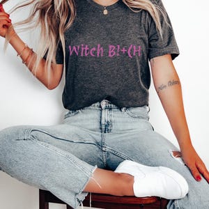 May include: A dark grey t-shirt with the text "Witch B!+CH" printed in pink on the front. The shirt is being worn by a person with long blonde hair and blue jeans.