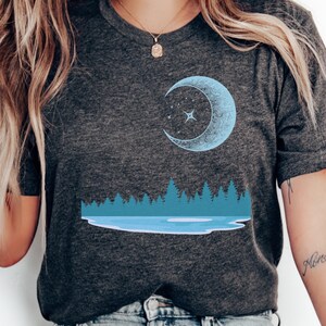 May include: Dark gray t-shirt featuring a light blue crescent moon and star design. Below the moon is a blue lake and forest scene. The shirt is a crew neck and has short sleeves.