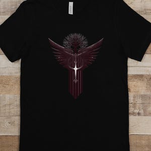 May include: Black t-shirt featuring a burgundy graphic of a stylised tree with wings and a cross. The tree is surrounded by a circle of leaves.