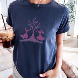 May include: A navy blue t-shirt featuring a pink graphic of two women dancing around a tree. The tree has pink leaves and roots.