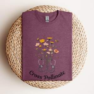 Könnte beinhalten: Ein lila T-Shirt mit einem Blumendesign und einem Stethoskop. Der Text "Cross Pollinate" ist auf dem Shirt aufgedruckt.