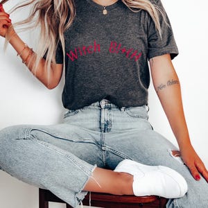 May include: A woman wearing a dark grey t-shirt with the text "Witch B*tch" in red lettering. She is wearing light blue jeans and white trainers.