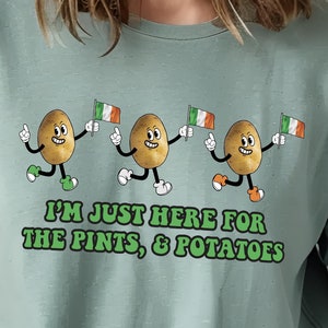 May include: A light green t-shirt with a graphic of three cartoon potatoes wearing green hats and holding Irish flags. The text "I'm just here for the pints & potatoes" is printed below the potatoes.