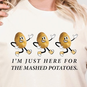 May include: Cream-colored t-shirt featuring a playful design of three animated potatoes with arms and legs, each with a smiling face. The text "I'M JUST HERE FOR THE MASHED POTATOES" is printed below the potatoes. The shirt is made of a soft material.