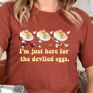 Puede incluir: Una mujer lleva una camiseta de color óxido con un gráfico de tres huevos rellenos y el texto "Solo estoy aquí por los huevos rellenos". Está sentada en un banco con una manta a cuadros y una almohada blanca.
