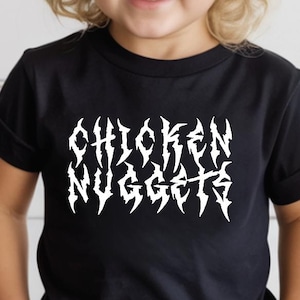 Może przedstawiać: Czarny t-shirt z białym napisem "CHICKEN NUGGETS" w stylizowanej czcionce. Dziecko ma na sobie musztardową czapkę i czarne okulary przeciwsłoneczne. Dziecko ma również na sobie czarno-białe szorty w kratę.
