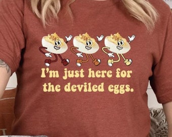Pode incluir: Uma mulher vestindo uma camiseta cor ferrugem com um gráfico de três ovos diabólicos de desenho animado. A camiseta diz "Estou aqui apenas para os ovos diabólicos."