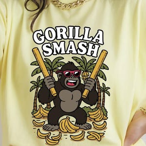 May include: Yellow t-shirt with the text "GORILLA SMASH" and a cartoon gorilla wearing sunglasses and holding two bamboo sticks. The gorilla is surrounded by bananas and palm trees.