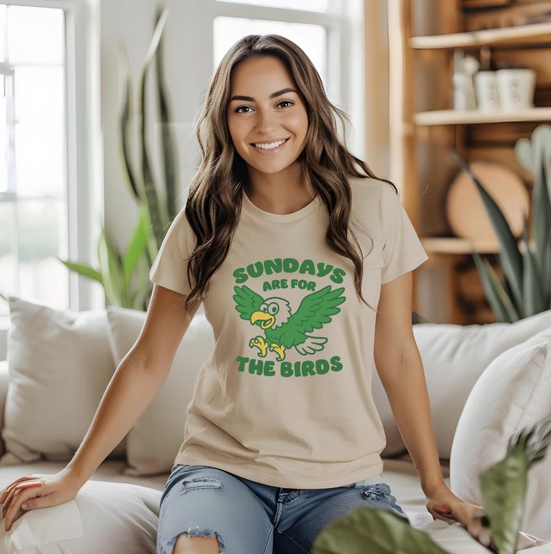 Op de afbeelding: Beige T-shirt met een groene en gele cartoon adelaar afbeelding. De tekst "SUNDAYS ARE FOR THE BIRDS" staat boven en onder de adelaar. Het shirt heeft korte mouwen en is gemaakt van een zacht materiaal.
