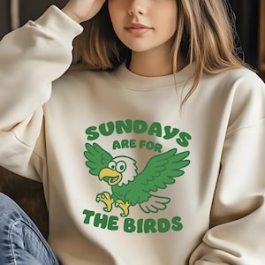 Op de afbeelding: Cr&egrave;mekleurige sweatshirt met een groene cartoon adelaar afbeelding en de tekst "SUNDAYS ARE FOR THE BIRDS". De adelaar heeft groene vleugels en een gele snavel en poten. De sweatshirt is een casual stijl.