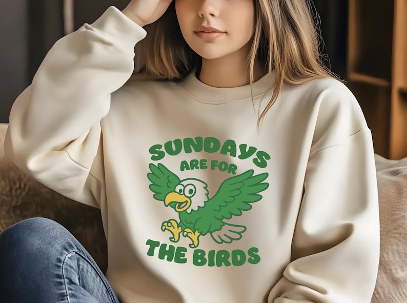 Op de afbeelding: Cr&egrave;mekleurige sweatshirt met een groene cartoon adelaar afbeelding en de tekst "SUNDAYS ARE FOR THE BIRDS". De adelaar heeft groene vleugels en een gele snavel en poten. De sweatshirt is een casual stijl.
