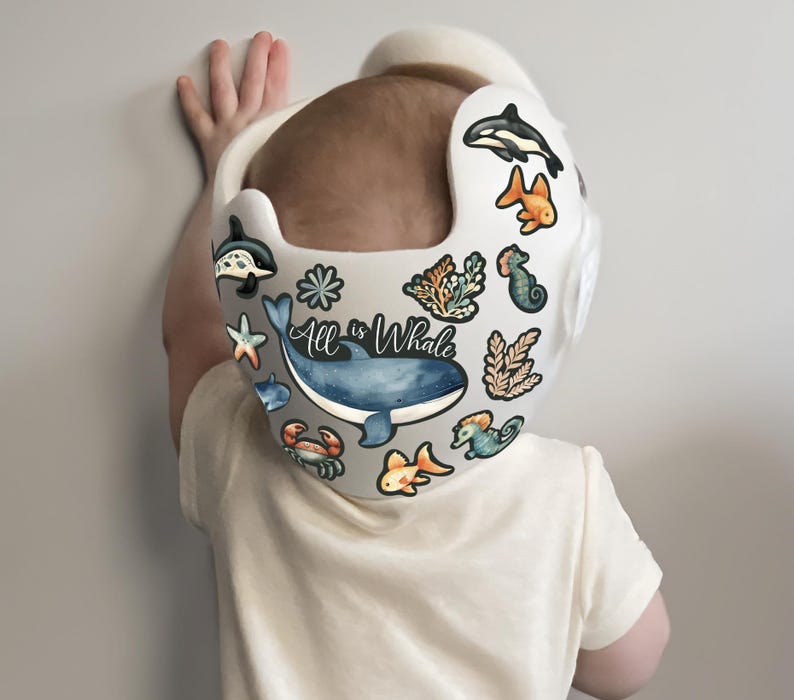 Op de afbeelding: Een witte helm met een blauwe walvis, een zeepaardje, een sterretje en andere stickers met een oceaan thema. De tekst "All is Whale" staat op de helm.