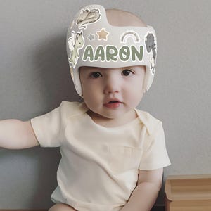 Puede incluir: Un beb&eacute; con un casco craneal gris claro con el nombre "AARON" en letras verdes. El casco est&aacute; decorado con pegatinas de animales de dibujos animados y arco&iacute;ris. El beb&eacute; lleva un body amarillo claro.