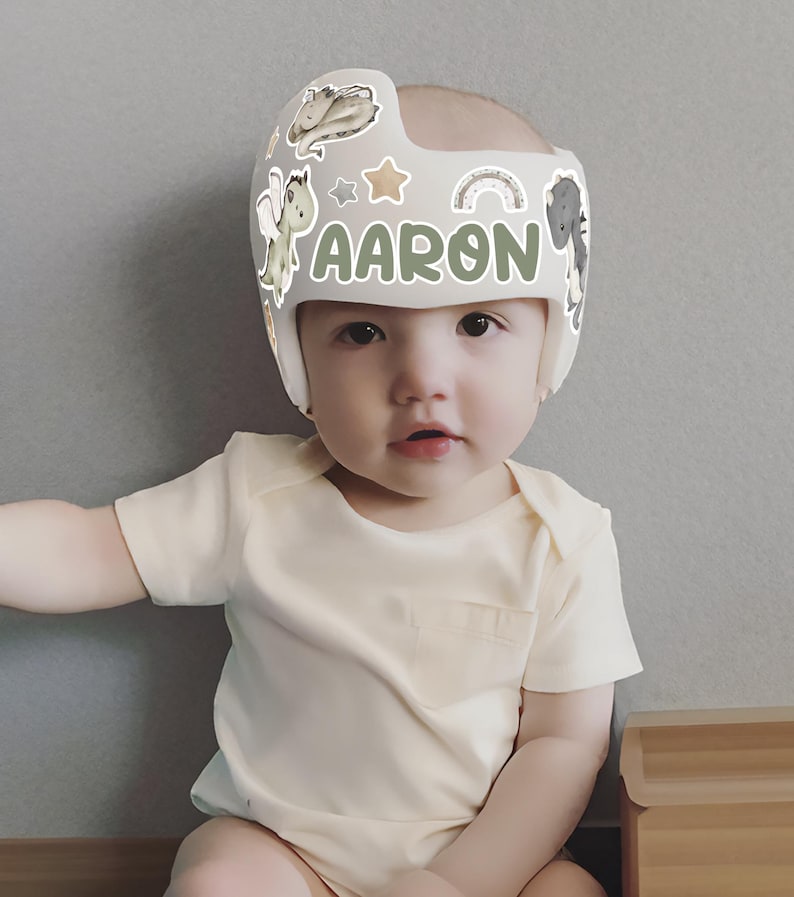 Puede incluir: Un beb&eacute; con un casco craneal gris claro con el nombre "AARON" en letras verdes. El casco est&aacute; decorado con pegatinas de animales de dibujos animados y arco&iacute;ris. El beb&eacute; lleva un body amarillo claro.