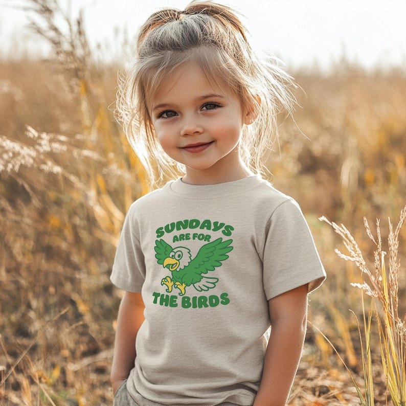 Op de afbeelding: Een lichtbeige t-shirt met de tekst "SUNDAYS ARE FOR THE BIRDS" en een groene adelaarsafbeelding. Het t-shirt wordt gedragen door een jong kind, staand in een veld met hoog gras.