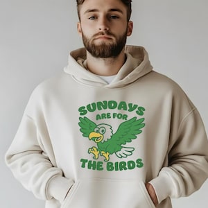 Op de afbeelding: Cr&egrave;mekleurige hoodie met een grafische print op de voorkant. De print toont een cartoon adelaar met groene vleugels en de tekst "SUNDAYS ARE FOR THE BIRDS" in het groen. De hoodie heeft een kangoeroezak en een capuchon met trekkoord.