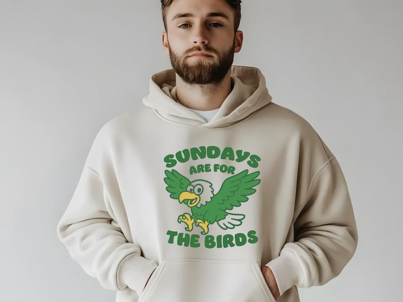 Op de afbeelding: Cr&egrave;mekleurige hoodie met een grafische print op de voorkant. De print toont een cartoon adelaar met groene vleugels en de tekst "SUNDAYS ARE FOR THE BIRDS" in het groen. De hoodie heeft een kangoeroezak en een capuchon met trekkoord.