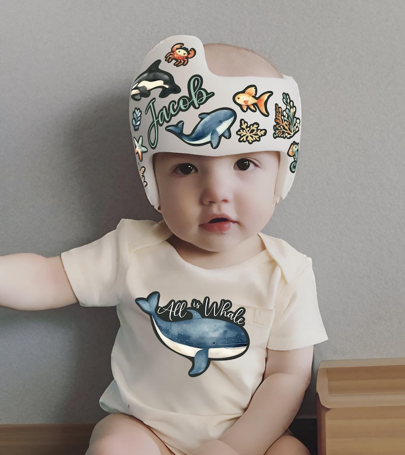 Op de afbeelding: Een baby draagt een witte helm met een blauwe walvis, een oranje vis, een sterretje en andere zeedieren. Op de helm staat de naam "Jacob". De baby draagt een witte romper met een blauwe walvis en de tekst "All is Whale".